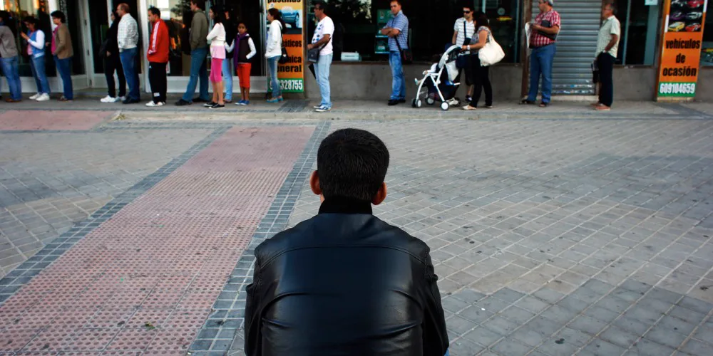 Bir meydanda sırtı dönük tek başına oturan genç; karşısında sıraya girmiş insanlar